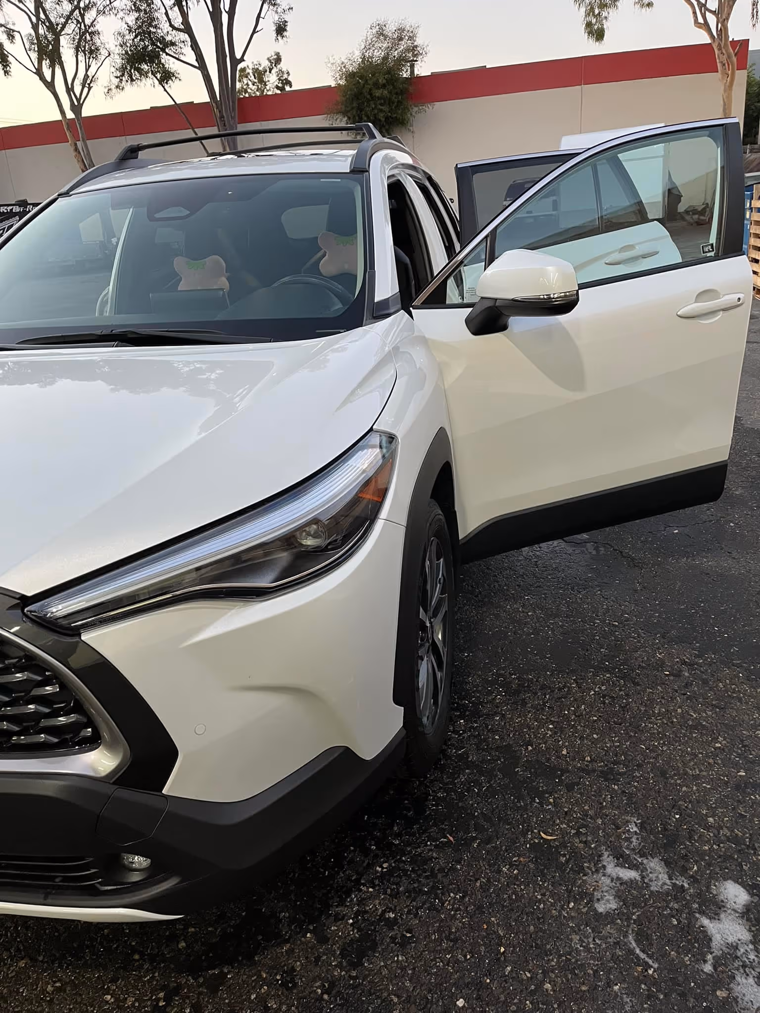 White car with the front passenger door open, parked on an asphalt surface near trees.