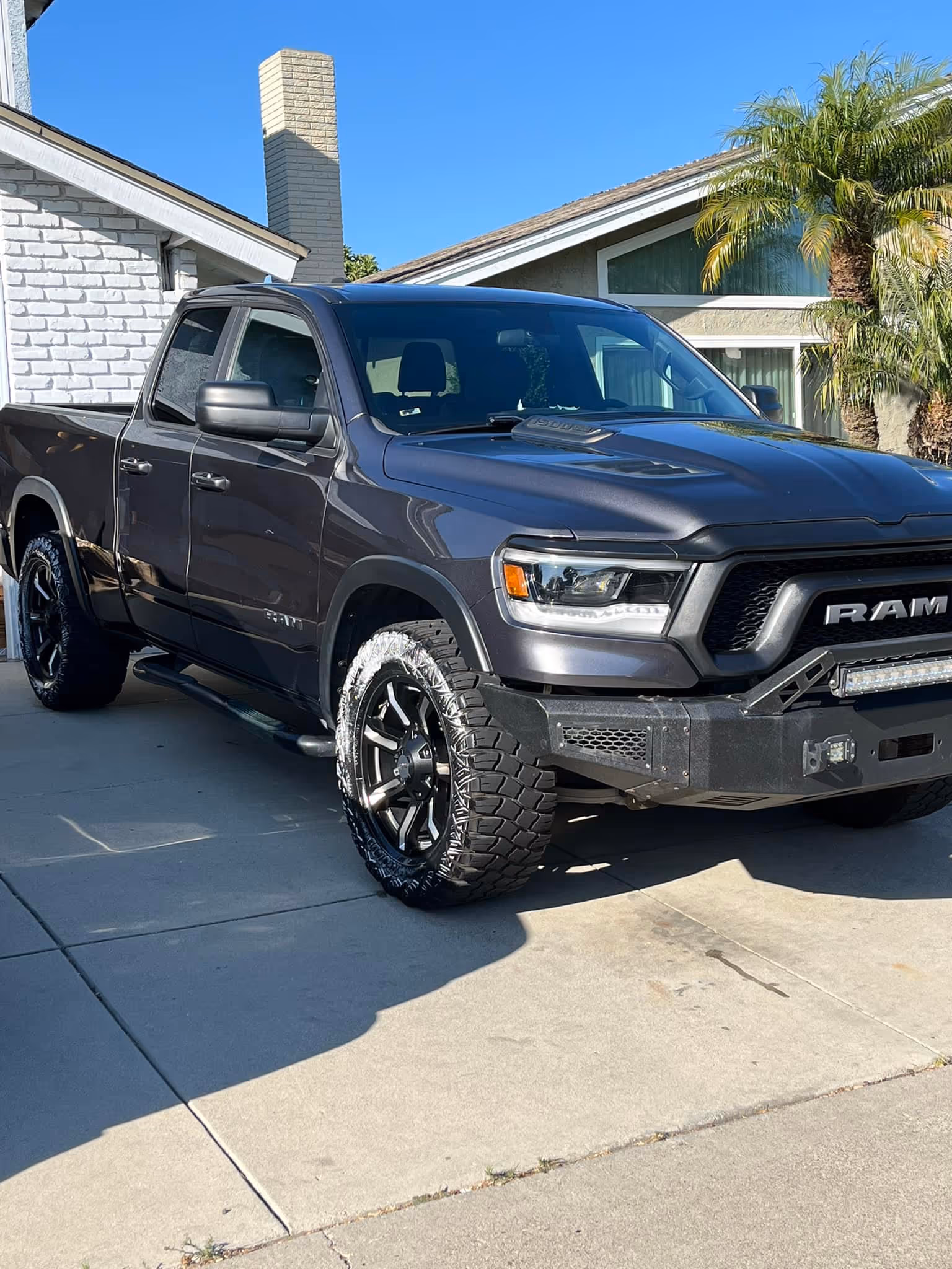 Black RAM pickup truck parked in driveway in front of house with palm trees and clear sky.
