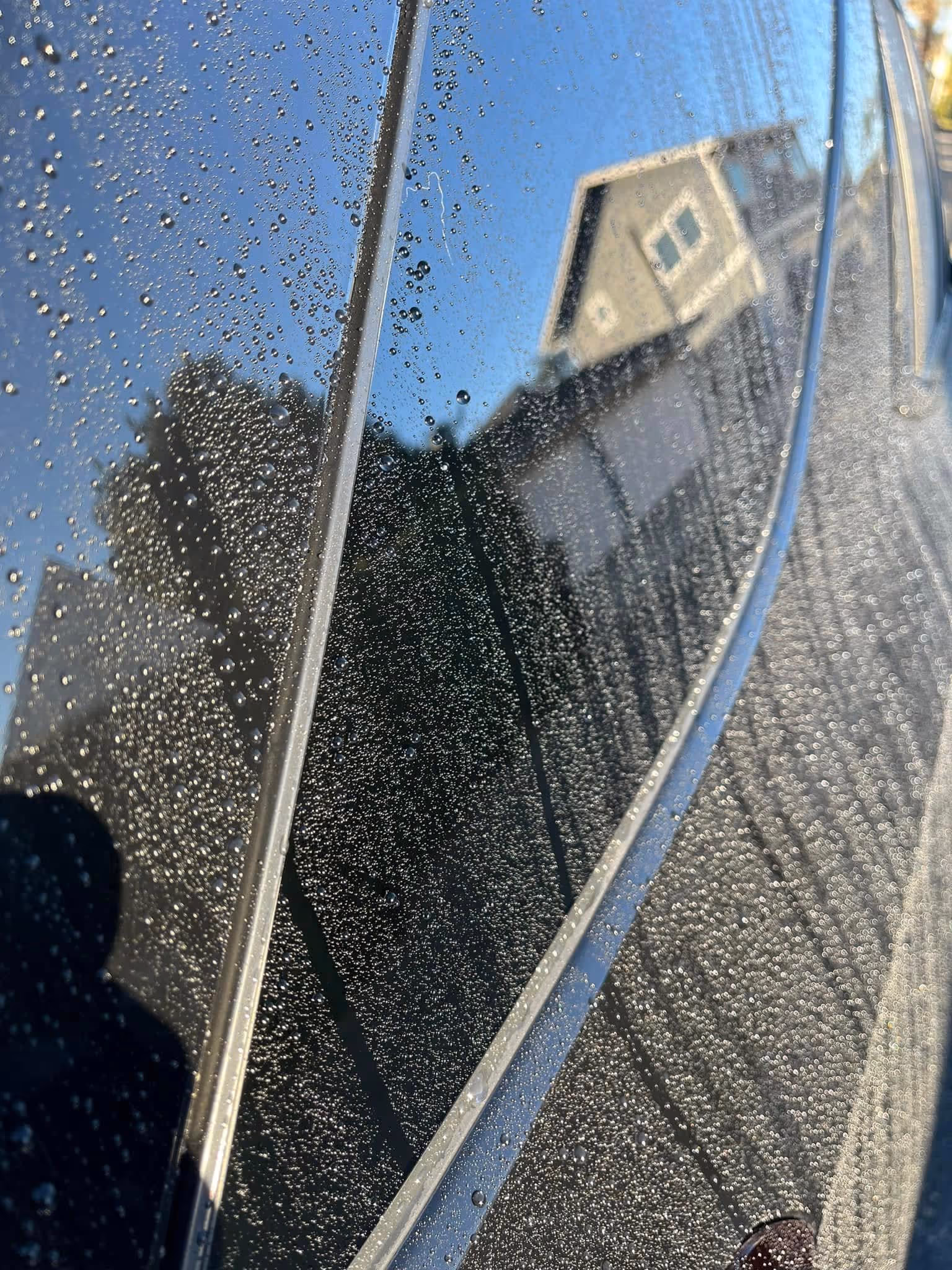 Close-up of water droplets on a shiny surface reflecting a house and trees under a clear blue sky.
