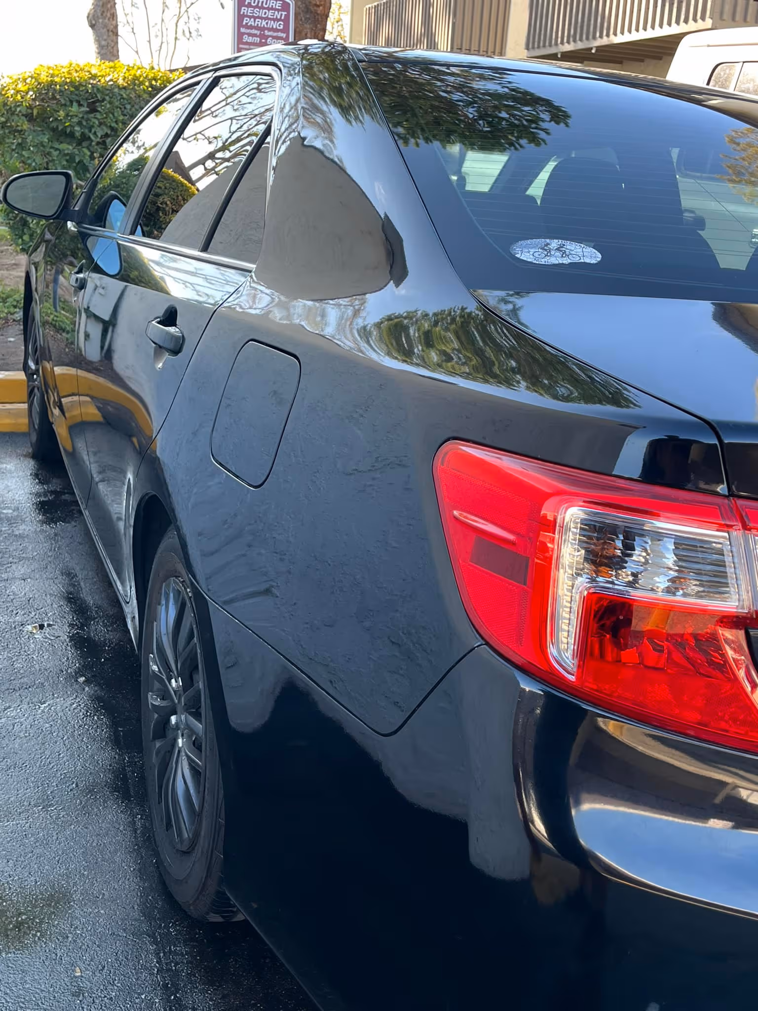 Black sedan parked next to yellow curb with reflections of trees and buildings on its surface.