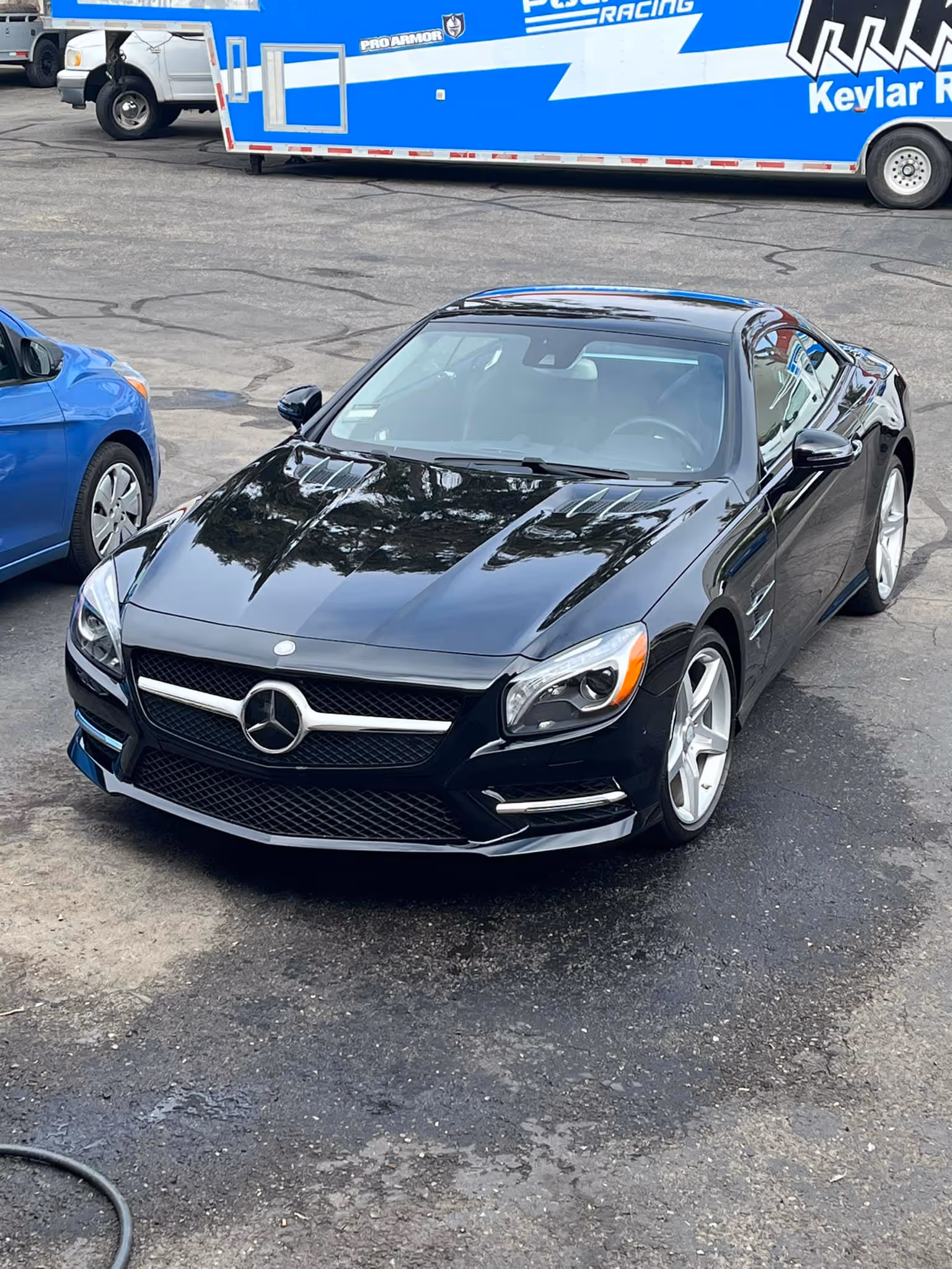 Black Mercedes-Benz sports car parked on asphalt with a blue car and trailer in background.