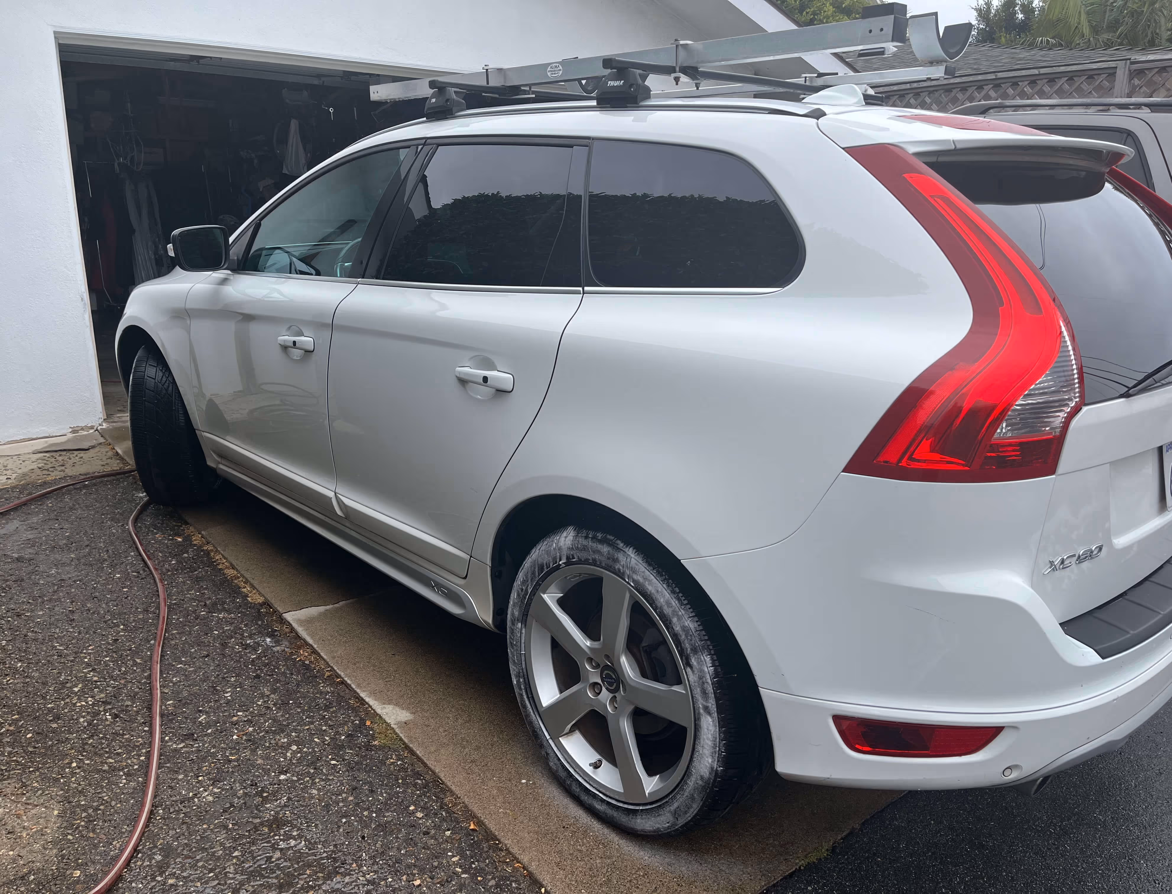 White Volvo XC60 SUV parked outside a garage with roof racks and a garden hose on the ground.